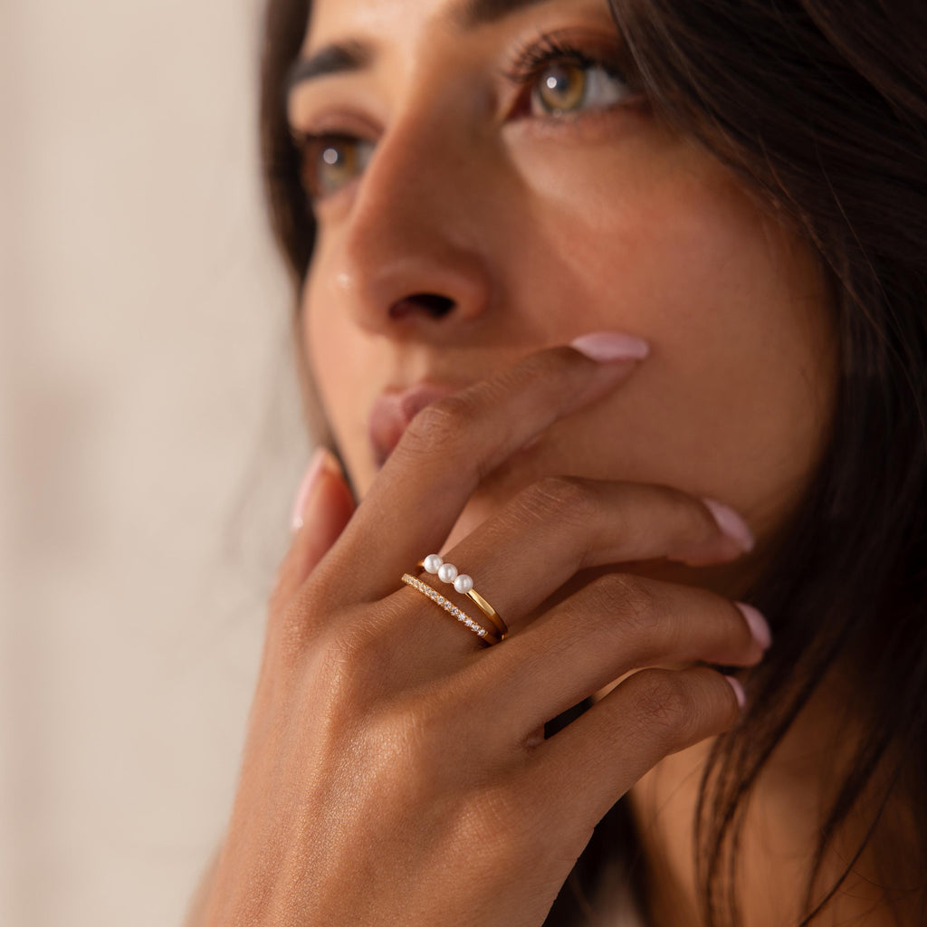 Woman wearing a gold Duo ring featuring three small pearls paired with a slim gold band set with pavé crystals, her hand resting near her lips with a soft focus on her face.