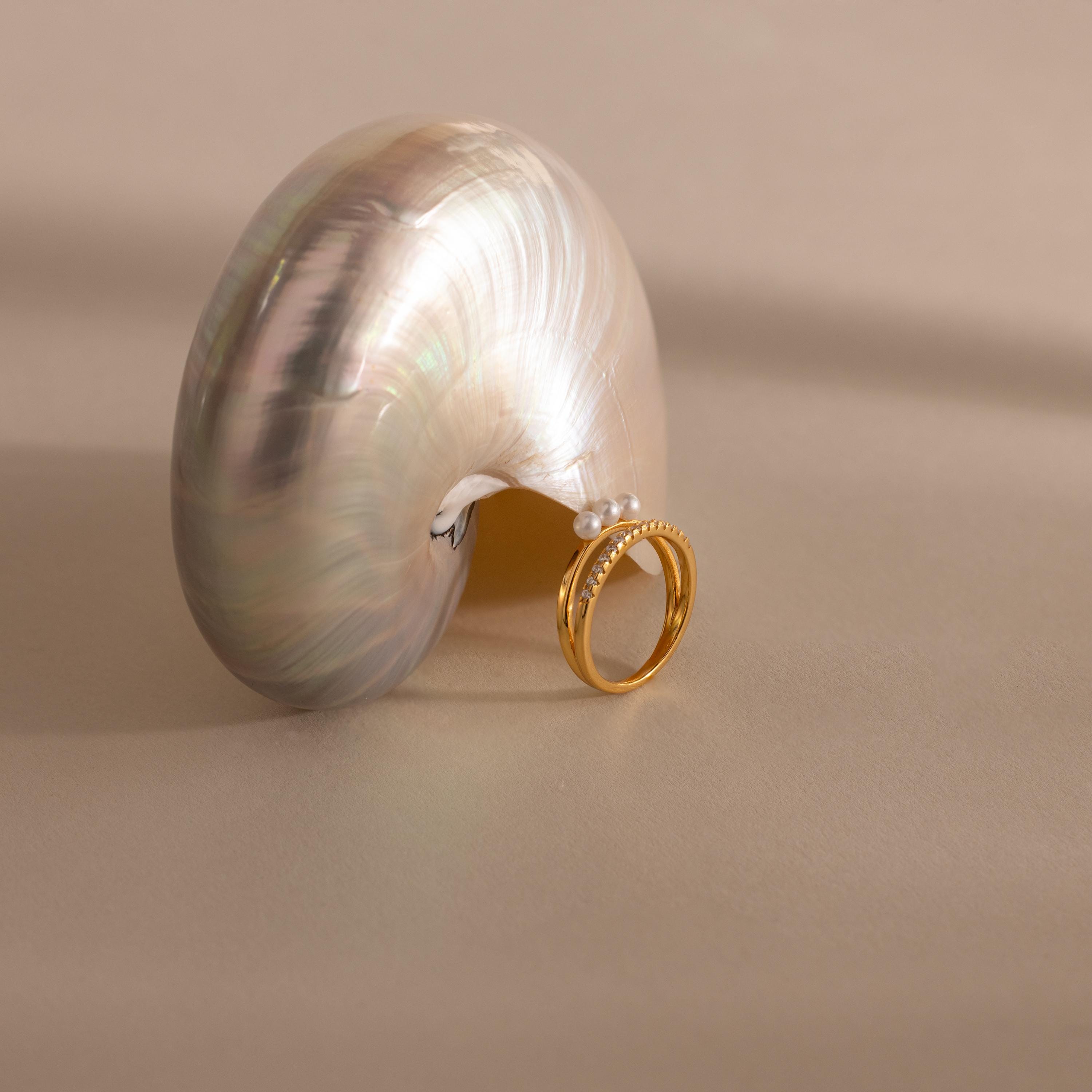 Gold duo ring with three small pearls paired with a slim pavé crystal band, displayed around the tip of a lustrous nautilus shell on a neutral background.