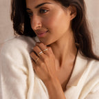 Woman in a soft cream sweater looking to the side, wearing a gold ring set with three pearls and a pavé crystal band, along with additional delicate gold rings.