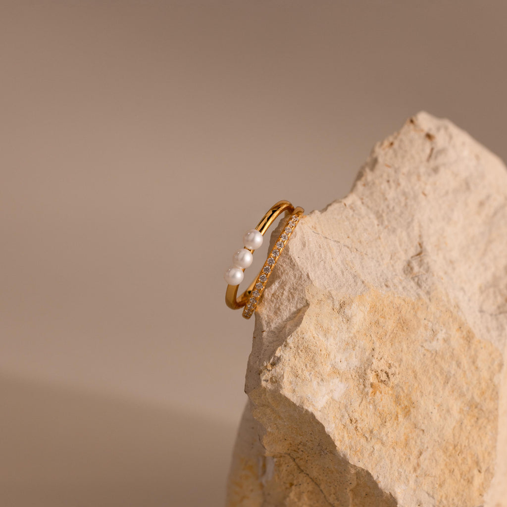 Gold Duo band ring featuring three small pearls on one band and a row of clear pavé crystals on the other, displayed balanced on a textured beige stone.