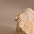 Gold Duo band ring featuring three small pearls on one band and a row of clear pavé crystals on the other, displayed balanced on a textured beige stone.