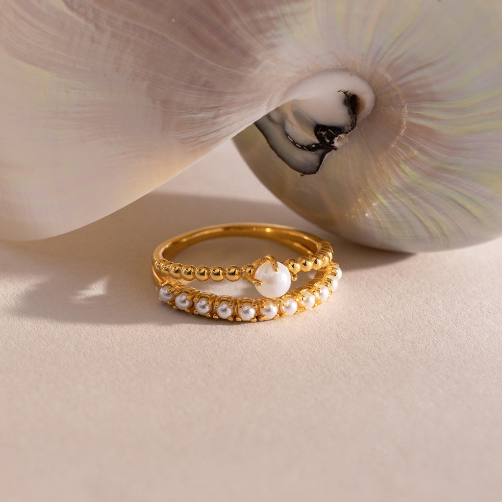 Gold ring set with beaded detailing, featuring a single central pearl on one band and a row of small pearls on the other, displayed beside a pearlescent seashell.