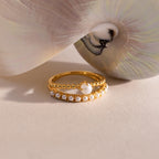 Gold ring set with beaded detailing, featuring a single central pearl on one band and a row of small pearls on the other, displayed beside a pearlescent seashell.