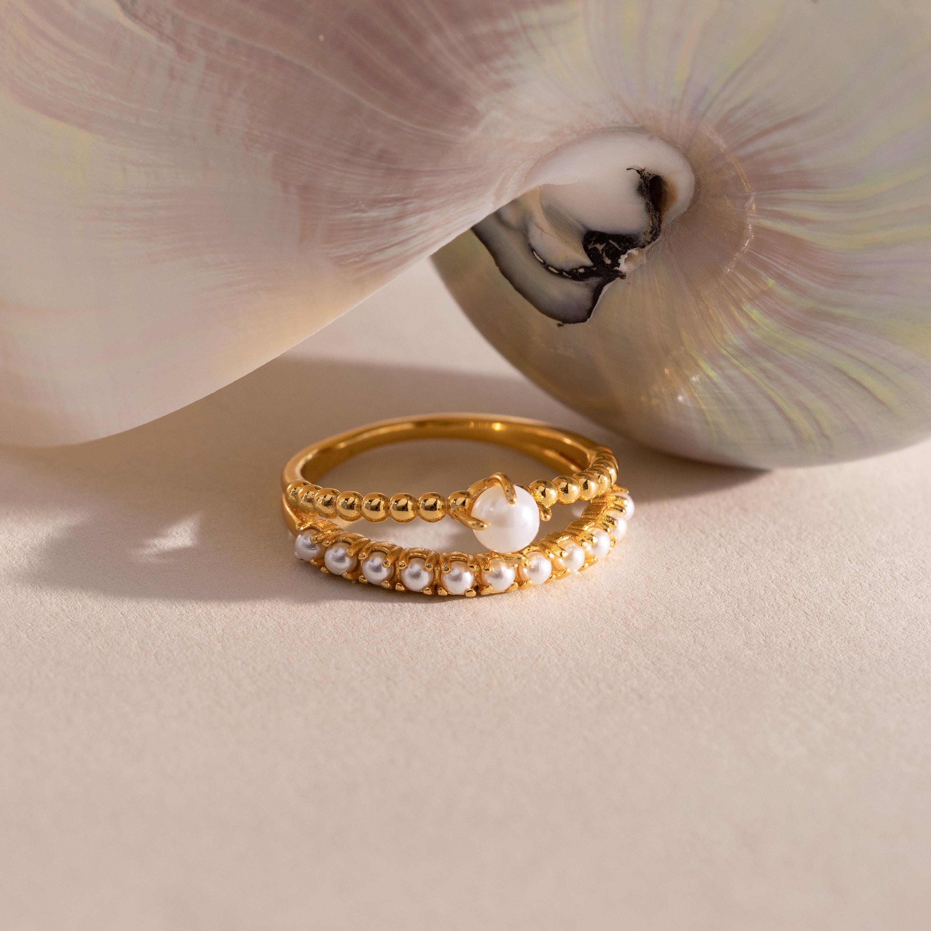 Gold ring set with beaded detailing, featuring a single central pearl on one band and a row of small pearls on the other, displayed beside a pearlescent seashell.