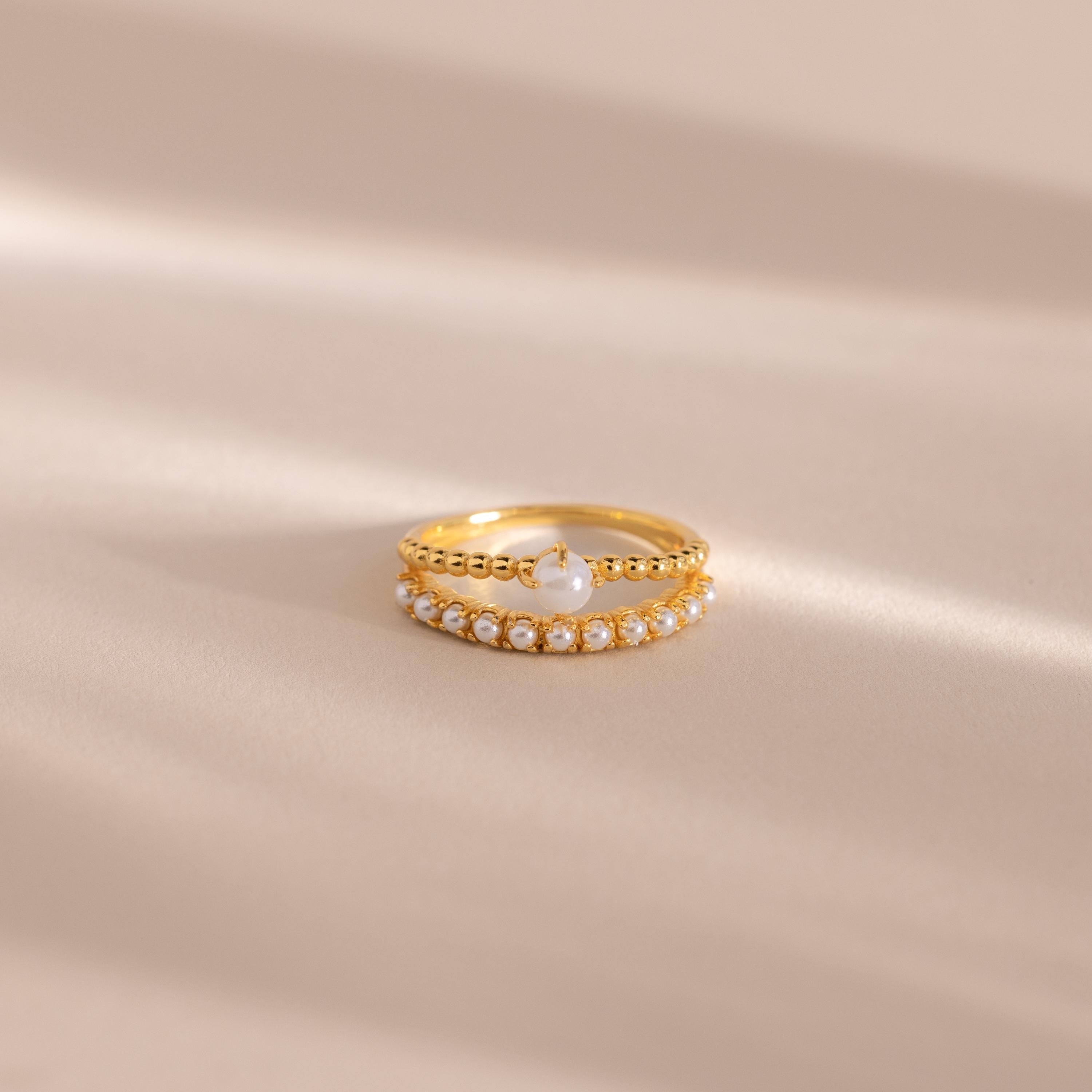 Gold beaded ring set featuring one band with a single central pearl and another adorned with a row of small pearls, displayed on a neutral beige surface with soft light shadows.