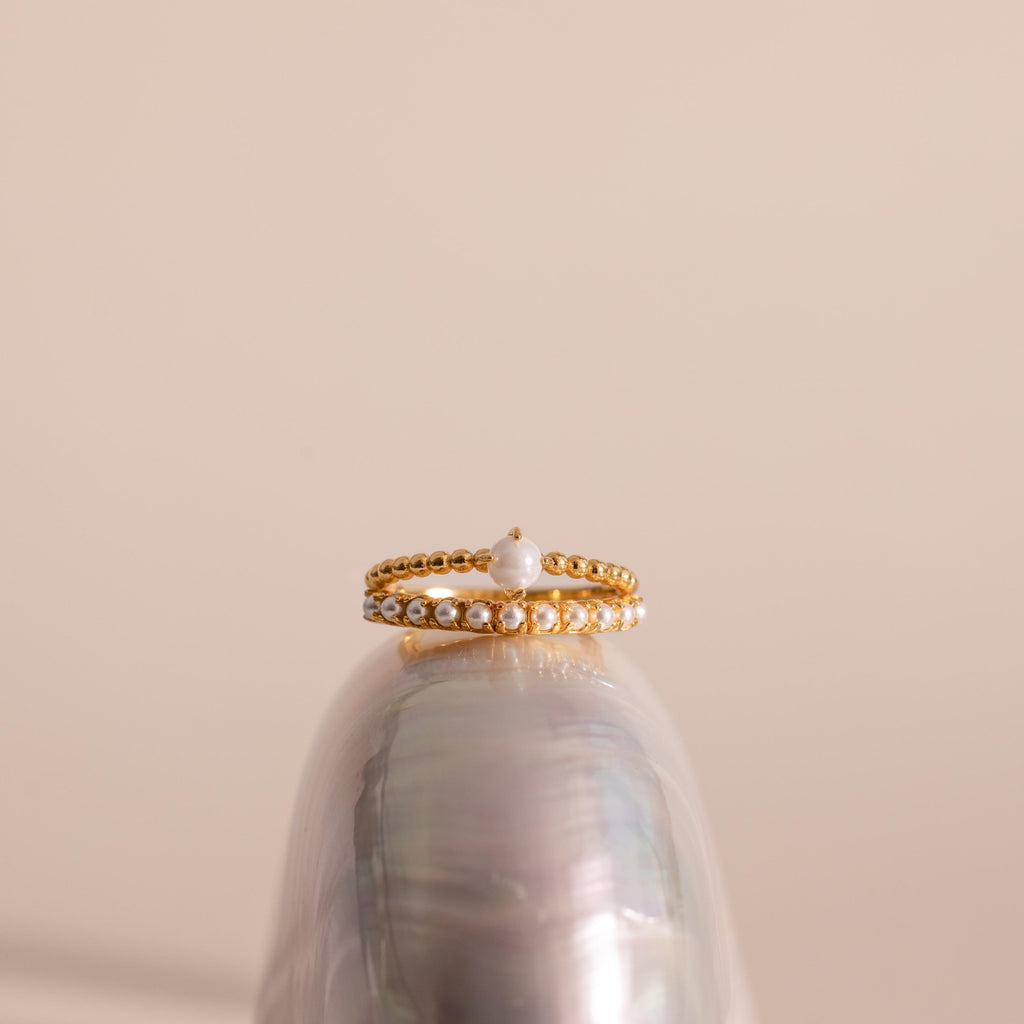 Gold beaded ring set featuring a single central pearl on one band and a row of small pearls on the other, displayed balanced atop a glossy seashell against a soft beige background.