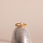 Gold beaded ring set featuring a single central pearl on one band and a row of small pearls on the other, displayed balanced atop a glossy seashell against a soft beige background.