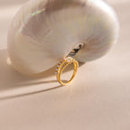 Gold beaded ring set with a central pearl and a lower band of small pearls, displayed leaning against a large iridescent seashell on a neutral surface.