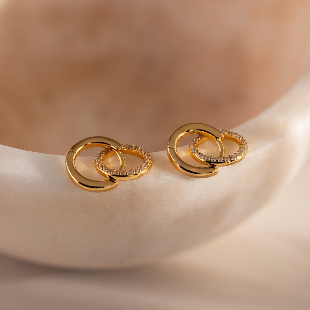 Pair of gold hoop earrings featuring two interlocking circles, one polished and one encrusted with small clear stones, displayed on a smooth, light toned stone surface.