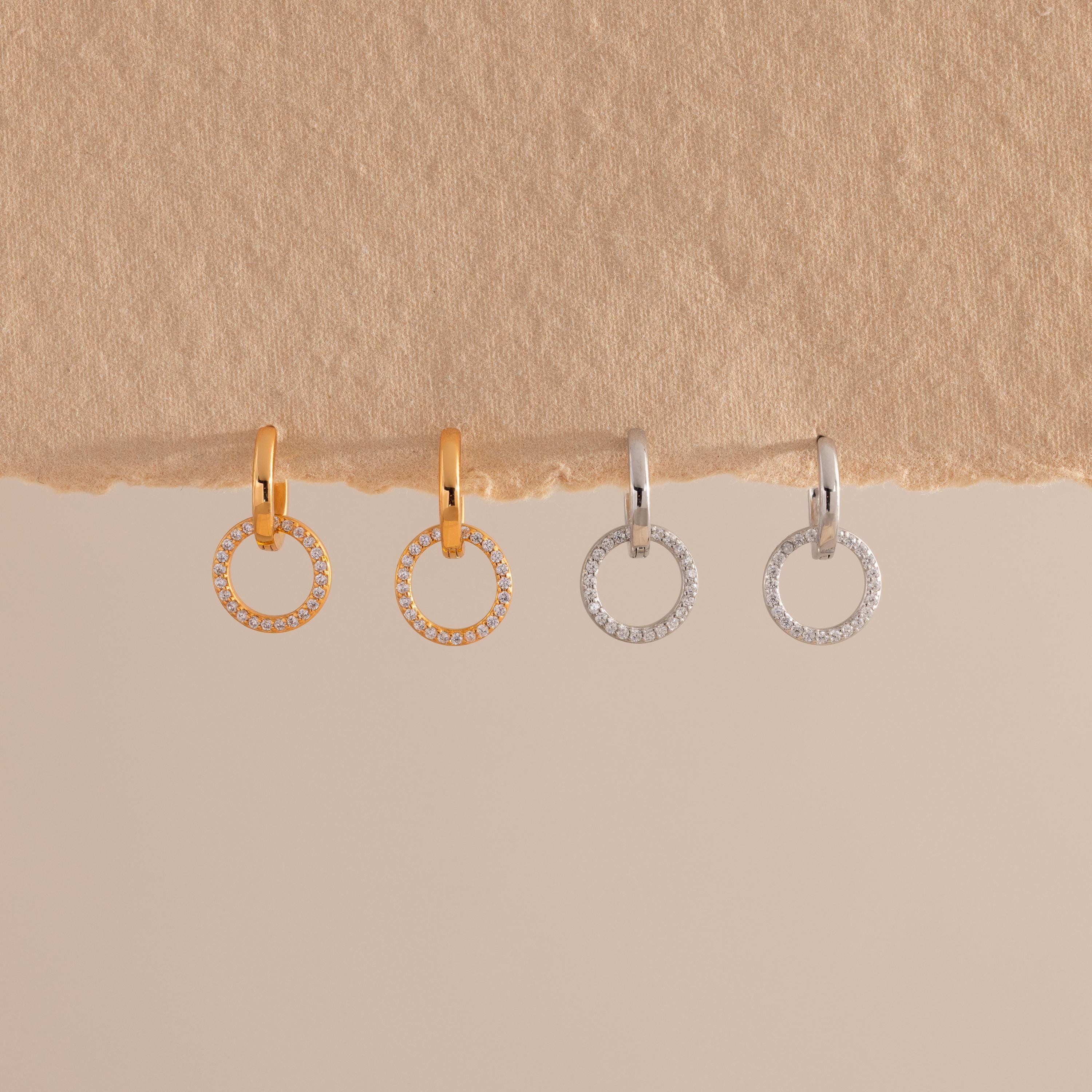 Two pairs of small hoop earrings with dangling open circles encrusted with clear stones, shown in gold and silver finishes, hanging against a textured beige surface.