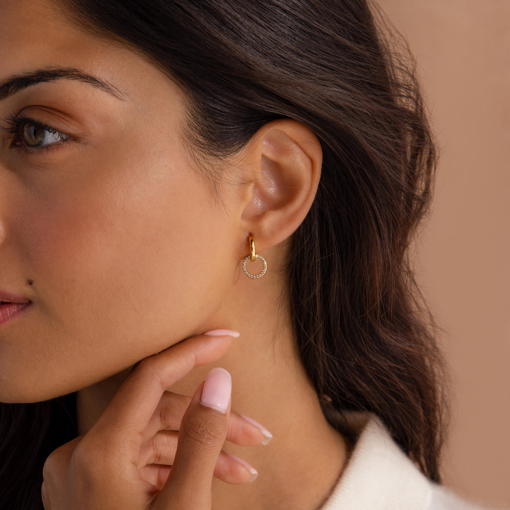 Woman wearing a small gold hoop earring with a dangling open circle encrusted with tiny clear stones, shown against a warm neutral background.