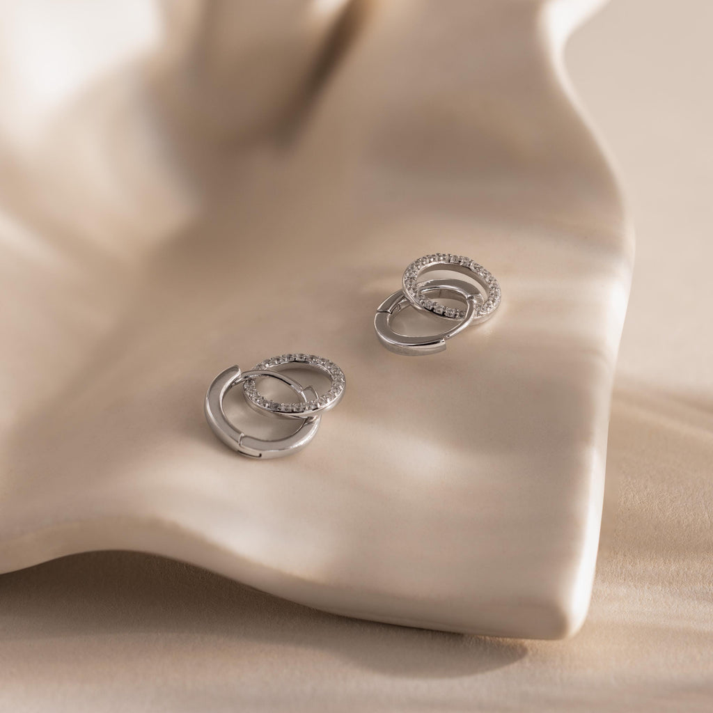 Pair of silver interlocking hoop earrings, one encrusted with small clear stones, displayed on a smooth, cream colored shell like surface.