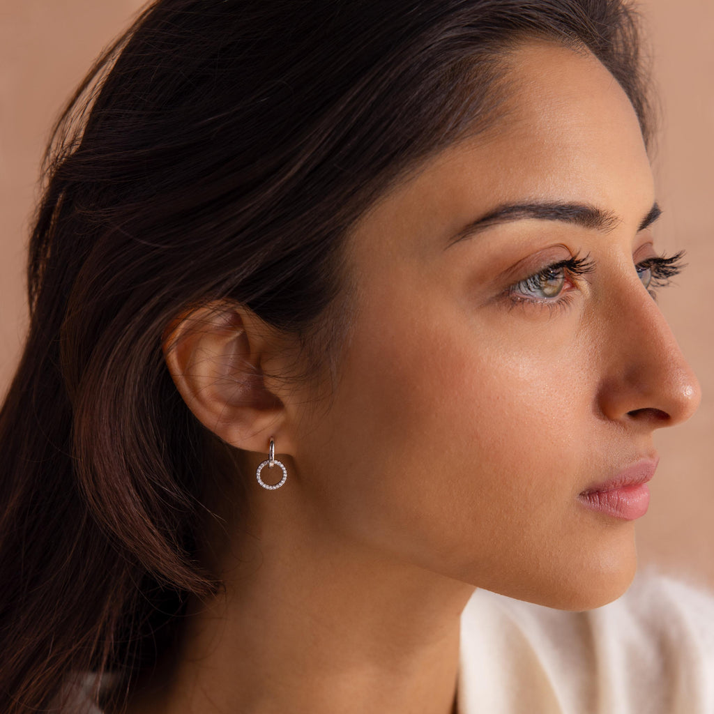 Woman wearing a small silver hoop earring with a dangling open circle encrusted with clear stones, shown in a close up side profile.