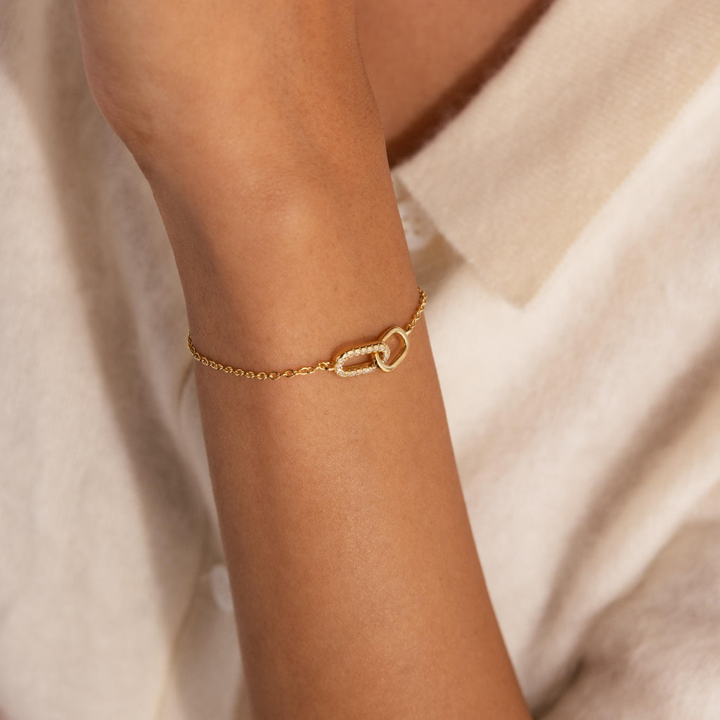 Gold chain bracelet with two interlocking oval links at the center, one link polished and the other encrusted with small clear stones, worn on the wrist.