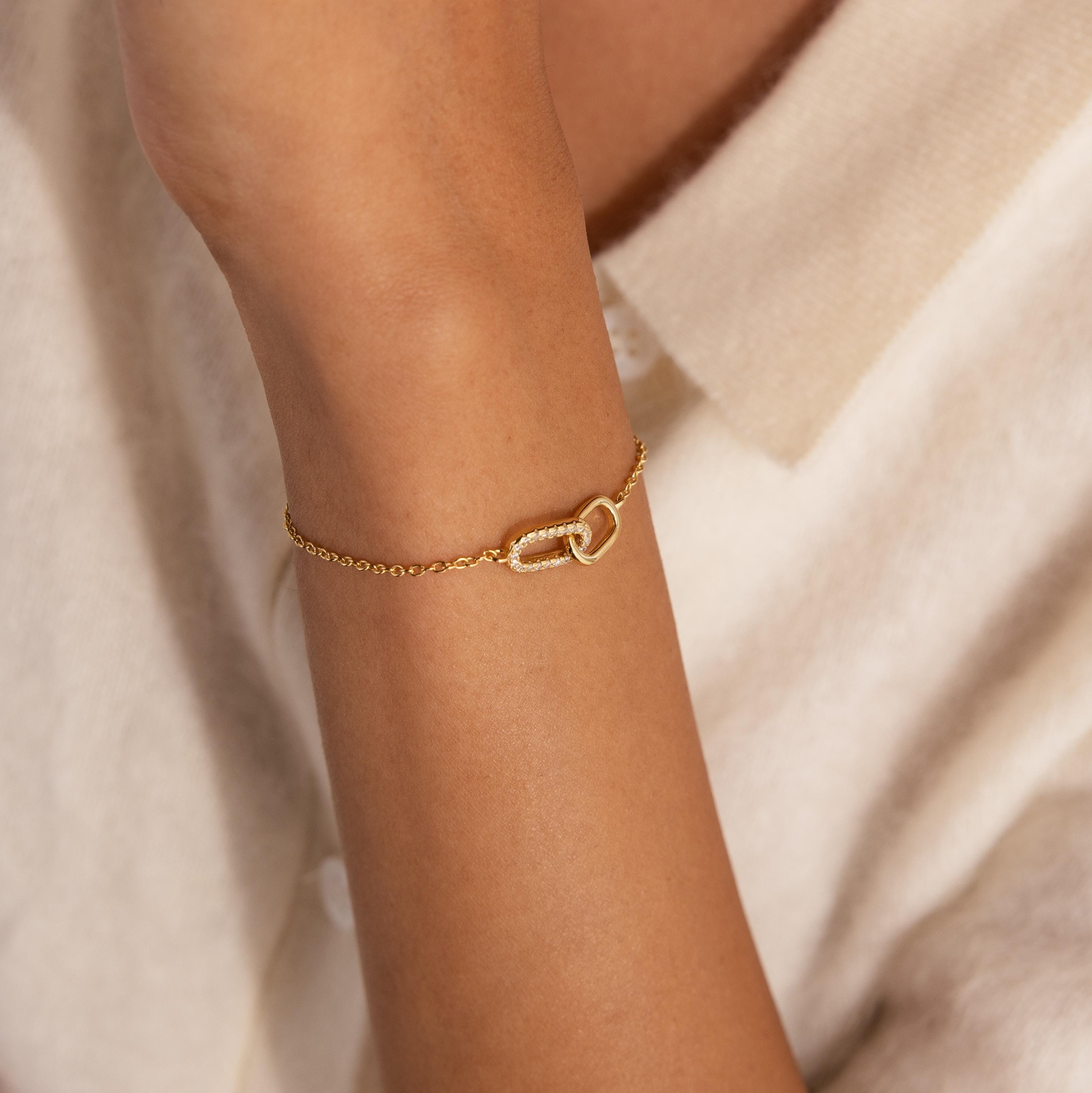 Gold chain bracelet with two interlocking oval links at the center, one link polished and the other encrusted with small clear stones, worn on the wrist.