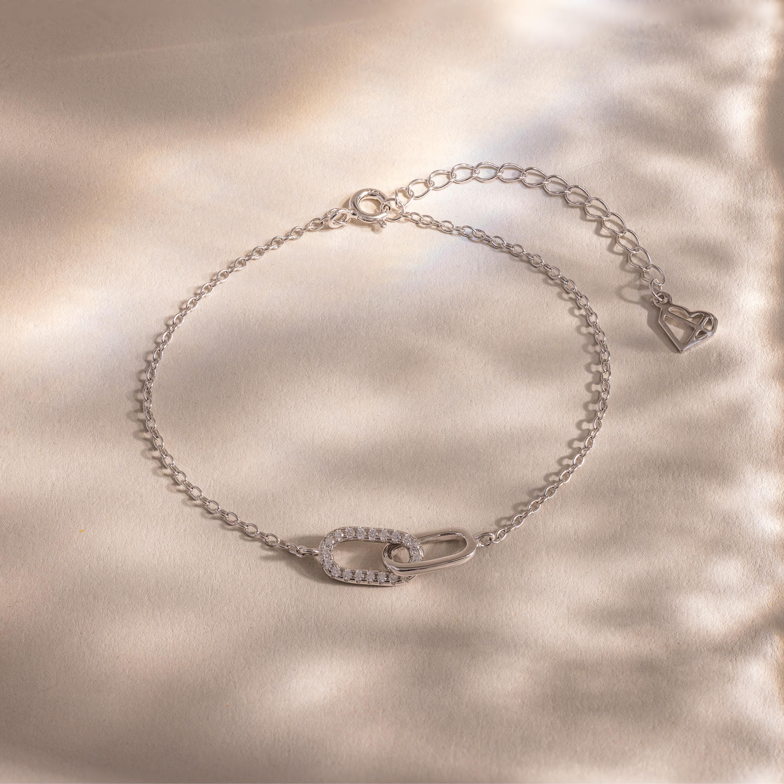 Delicate silver chain bracelet with two interlocking oval links at the center, one polished and one encrusted with small clear stones, displayed on a softly lit neutral background.