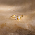 Gold ring with a emerald cut clear gemstone and a pear-shaped opal set side by side, displayed on a textured beige stone surface.