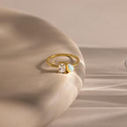 Gold ring with a slim band, featuring a rectangular clear gemstone and a pear-shaped opal side by side, displayed on a smooth, curved beige surface with soft shadows.