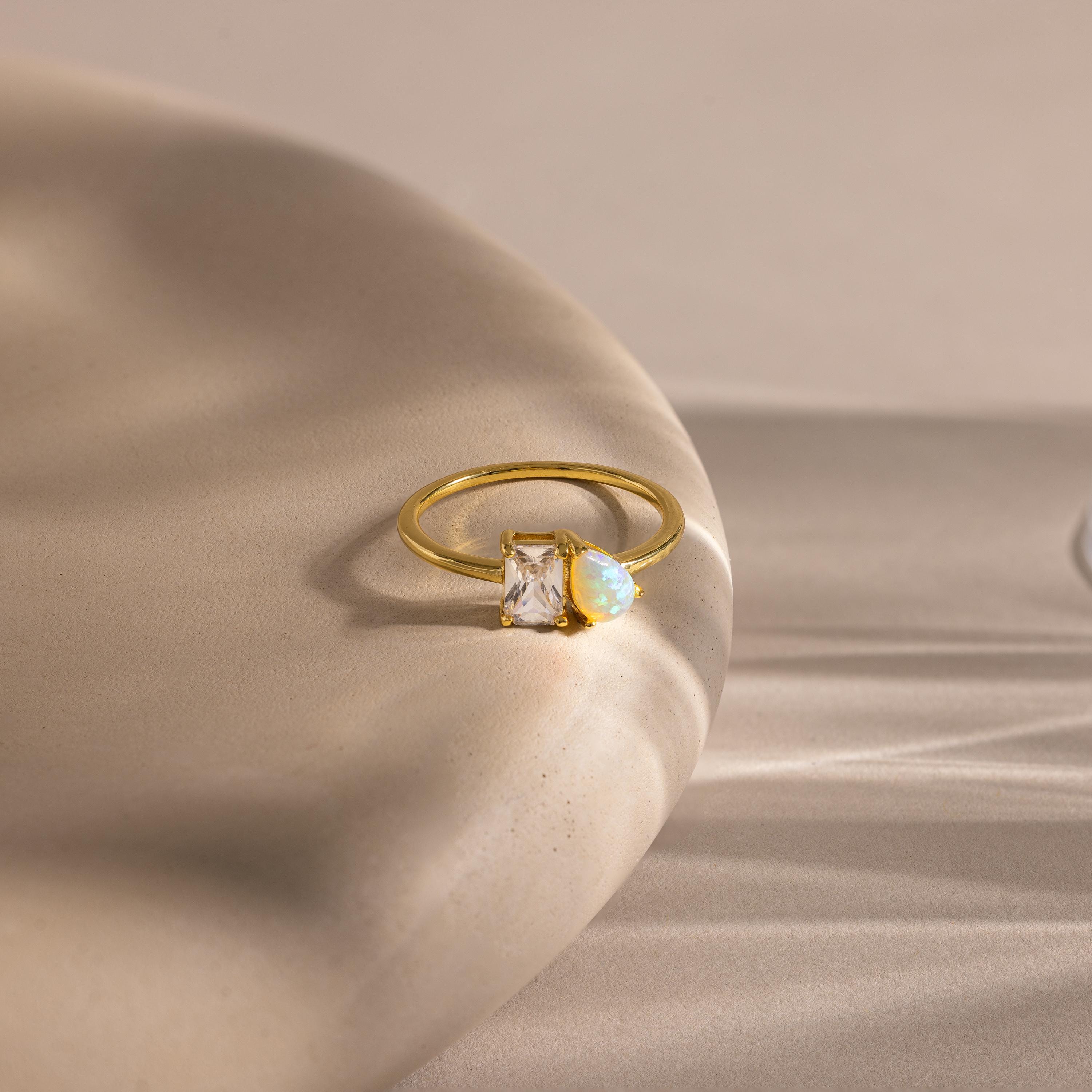 Gold ring with a slim band, featuring a rectangular clear gemstone and a pear-shaped opal side by side, displayed on a smooth, curved beige surface with soft shadows.