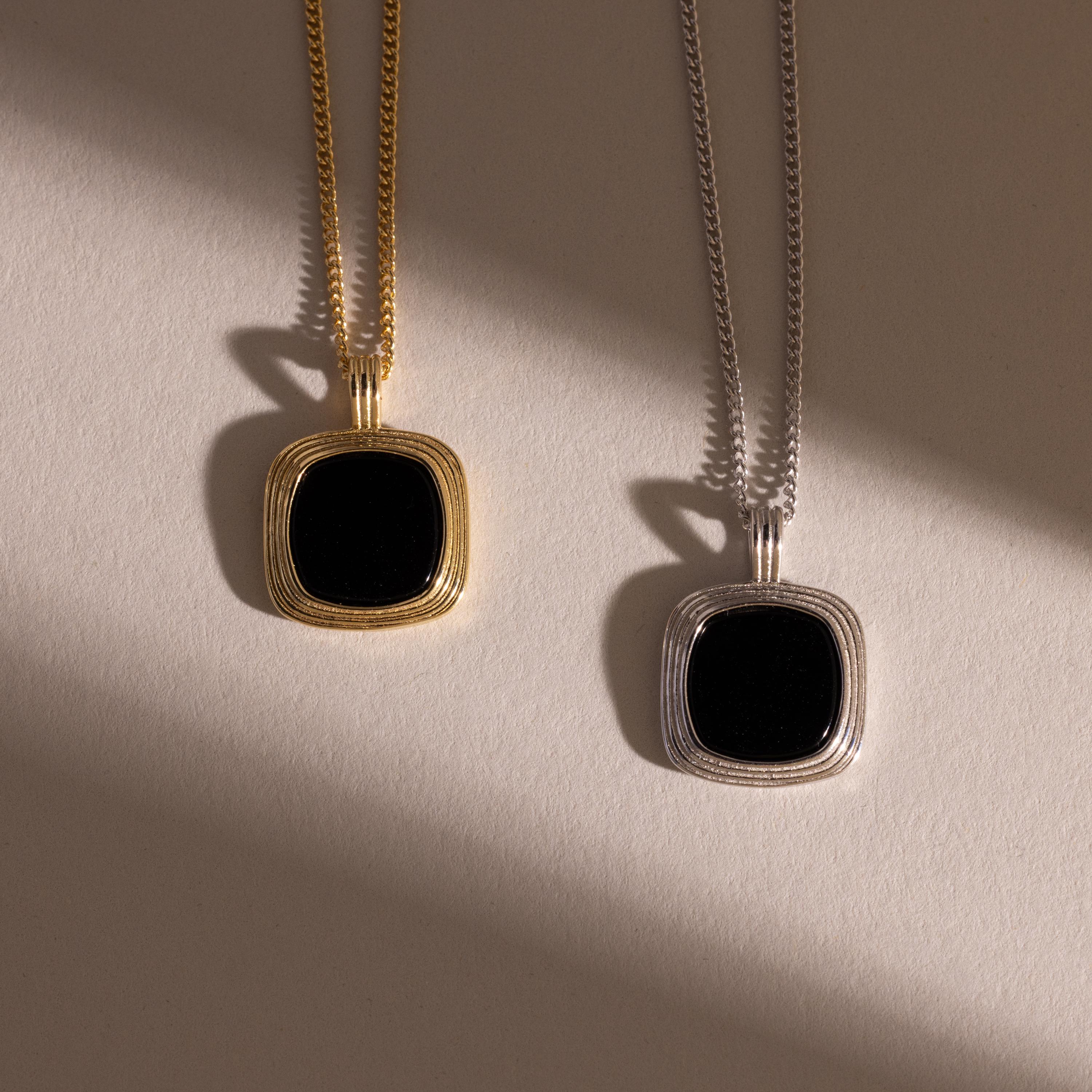 Two square black onyx pendant necklaces, one with a gold frame and chain and the other with a silver frame and chain, displayed side by side on a neutral surface with soft shadows.