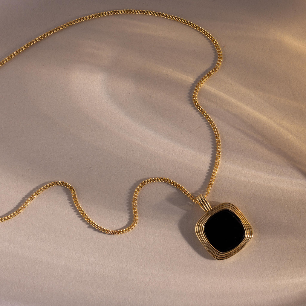 Gold necklace with a square black onyx pendant framed in layered gold detailing, displayed on a neutral surface with soft, warm light and shadows.
