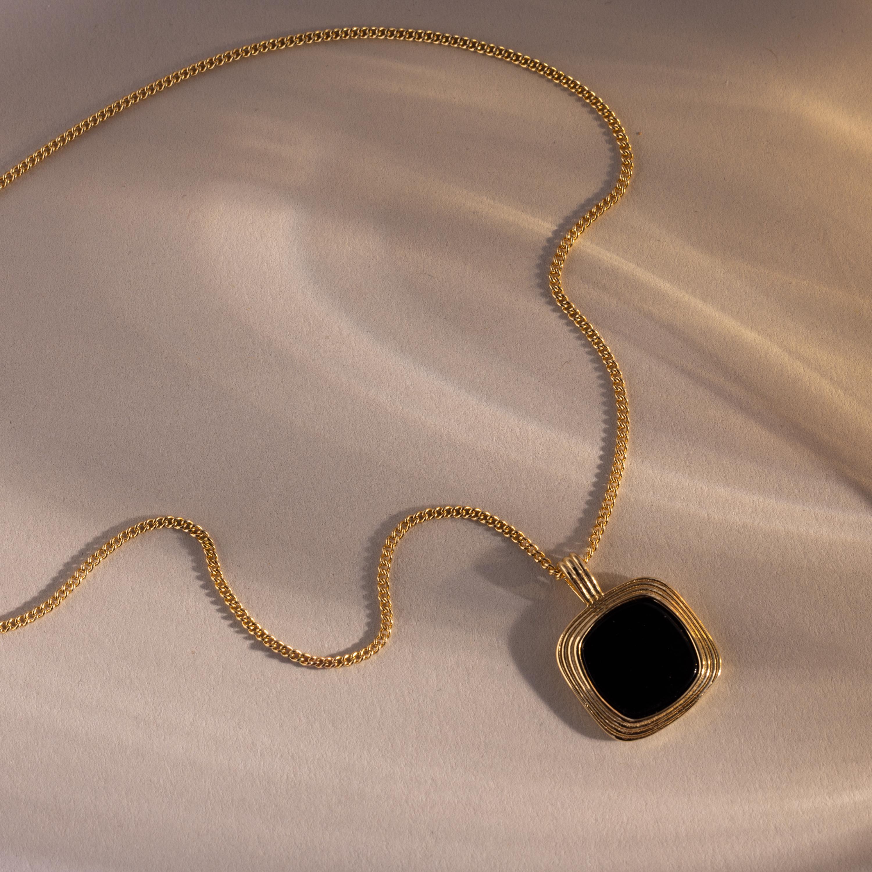Gold necklace with a square black onyx pendant framed in layered gold detailing, displayed on a neutral surface with soft, warm light and shadows.