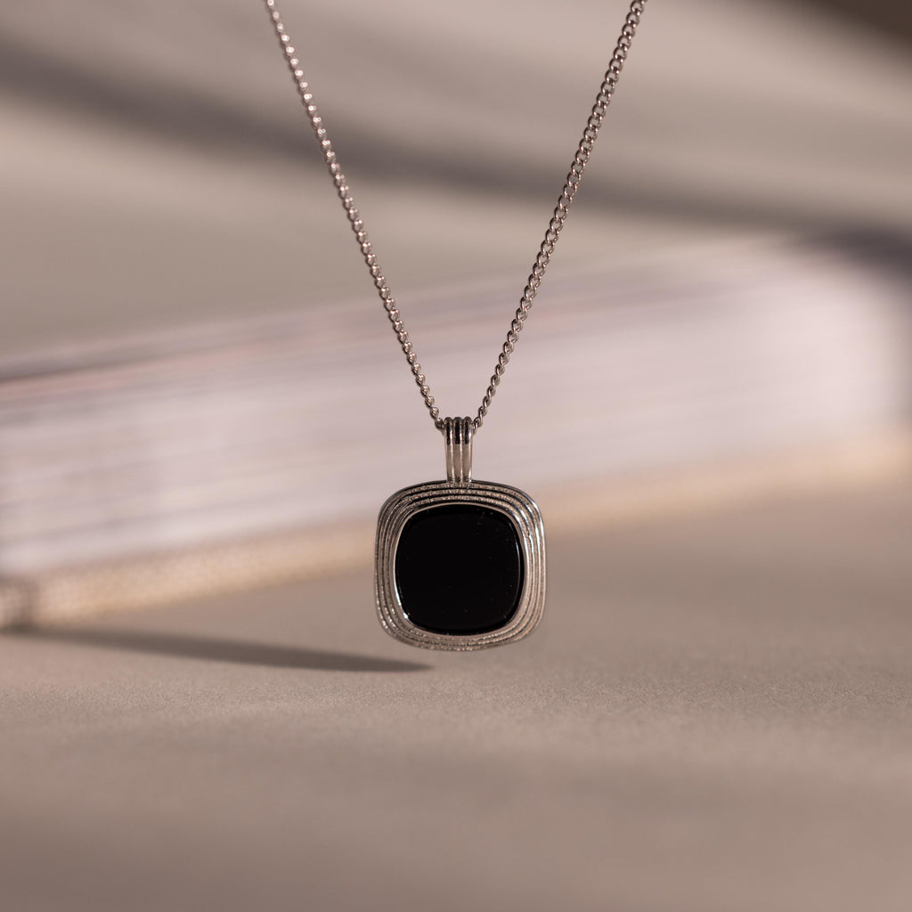 Silver chain necklace featuring a square black onyx pendant in a textured gold bezel, hanging against a neutral background with soft lighting.
