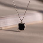 Silver chain necklace featuring a square black onyx pendant in a textured gold bezel, hanging against a neutral background with soft lighting.