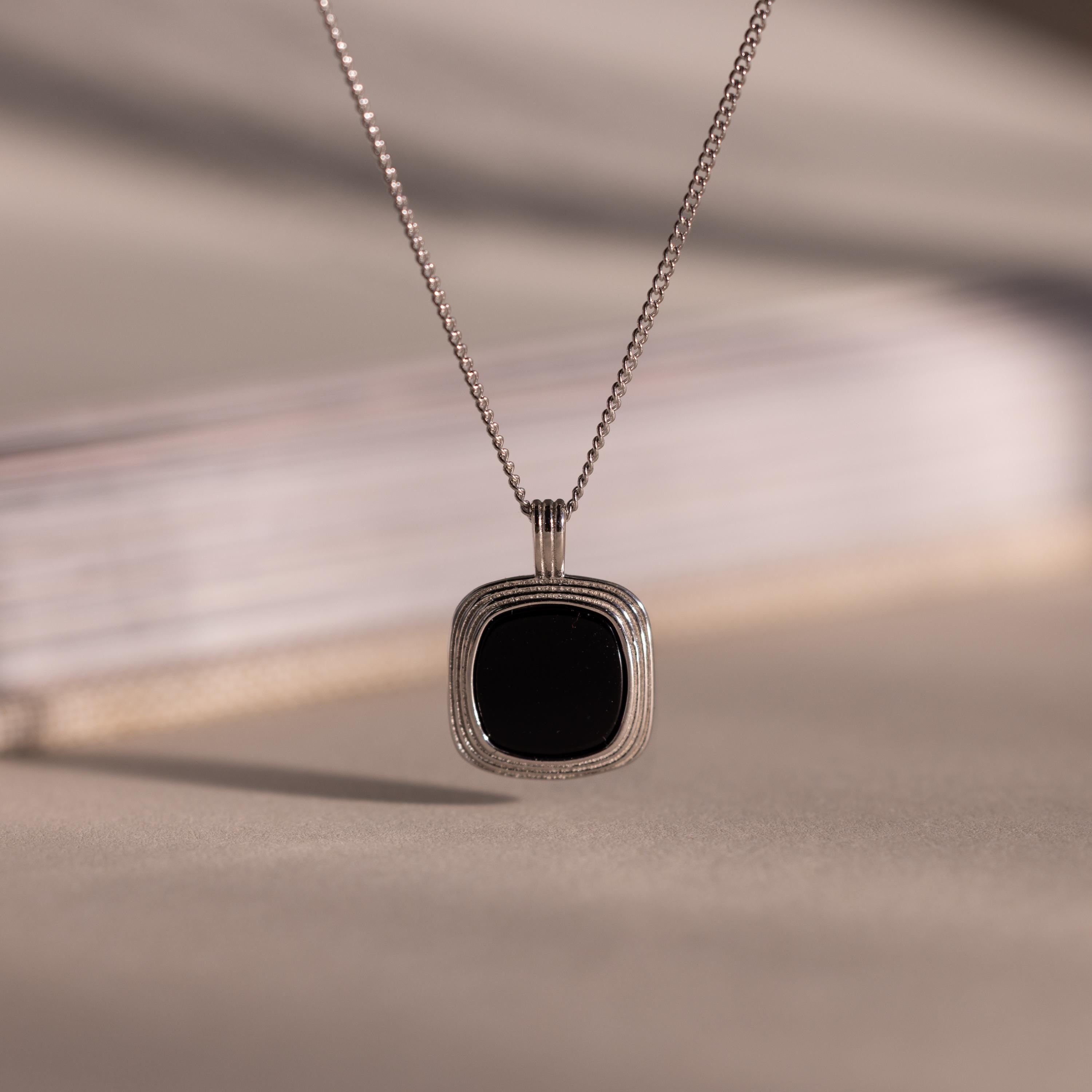 Silver chain necklace featuring a square black onyx pendant in a textured gold bezel, hanging against a neutral background with soft lighting.