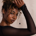 Woman in a sheer black long sleeve top holding a silver chain necklace with a square black onyx pendant in a textured silver bezel, gazing confidently at the camera.