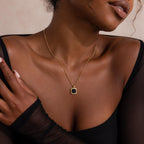 Woman wearing a thin gold chain necklace with a square black onyx pendant framed in gold, styled with a sheer black top.