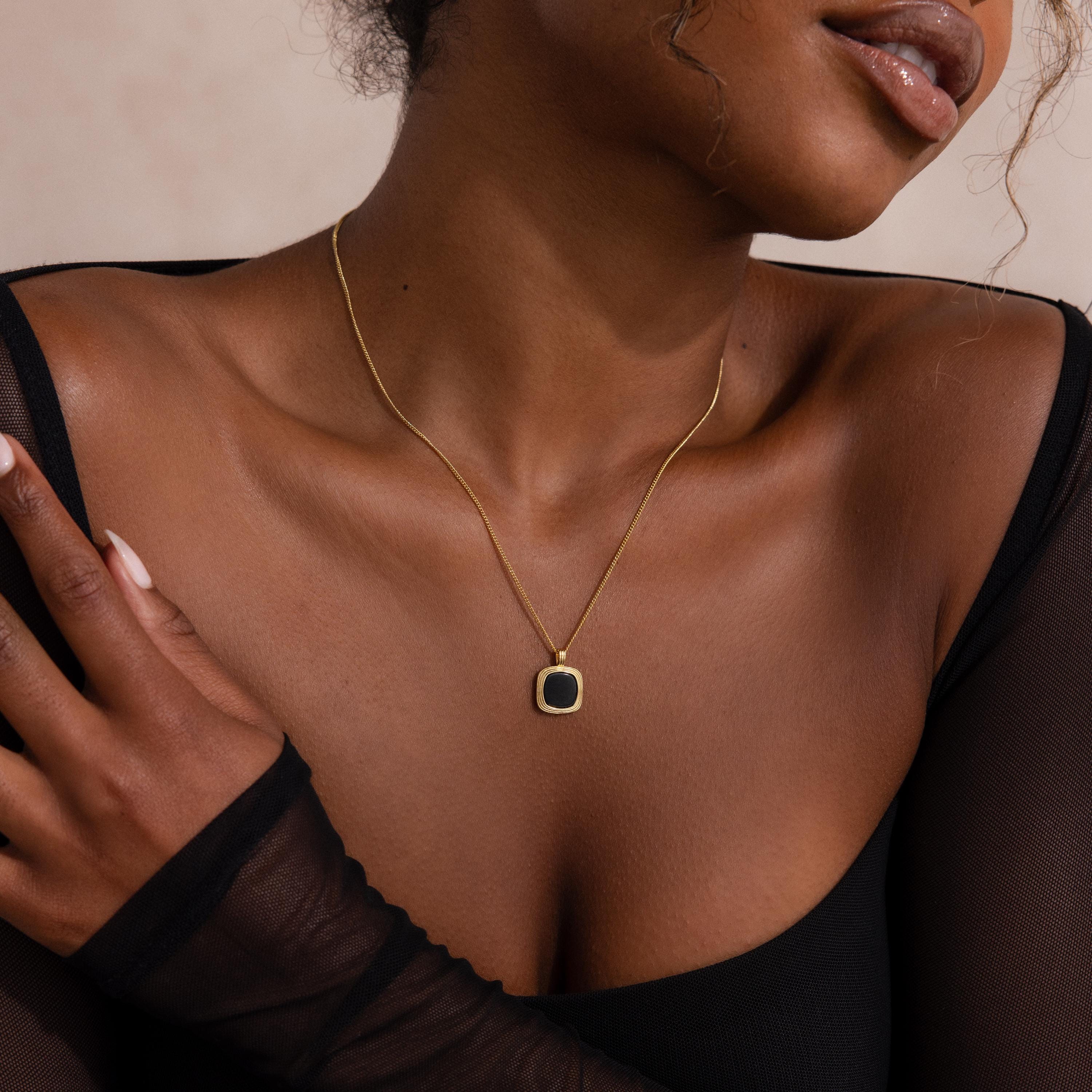 Woman wearing a thin gold chain necklace with a square black onyx pendant framed in gold, styled with a sheer black top.