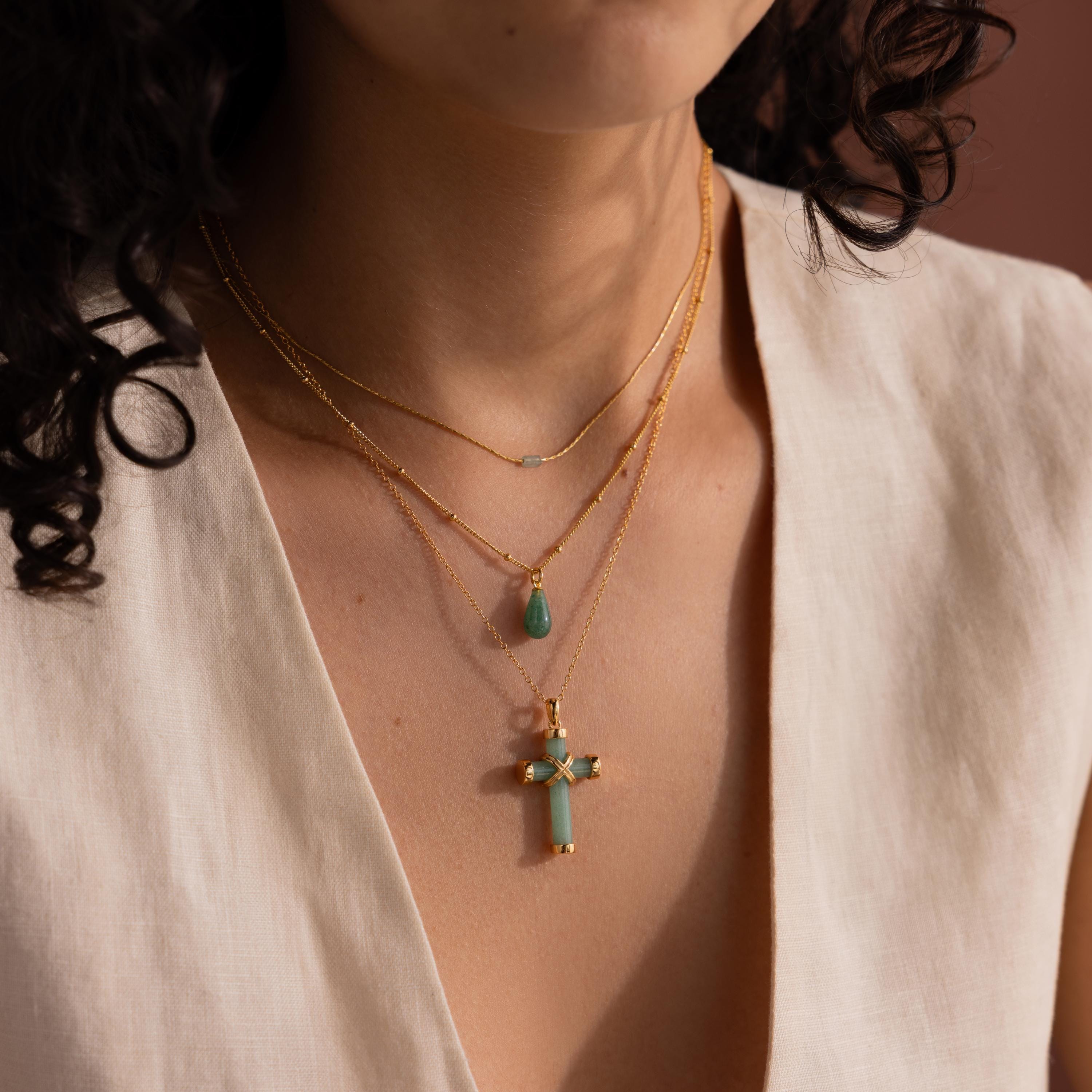 A woman styles the Jade Cross Necklace, featuring natural jade pendants, with other layered gold-tone necklaces and a V-neck beige top.
