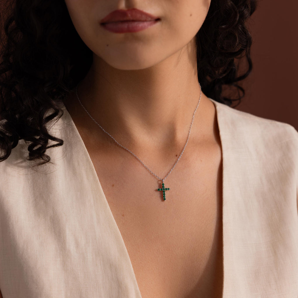 Woman wearing a silver chain necklace with a small cross pendant set with green gemstones, styled with a cream sleeveless V-neck top.