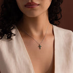 Woman wearing a silver chain necklace with a small cross pendant set with green gemstones, styled with a cream sleeveless V-neck top.