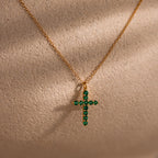 Gold chain necklace featuring a cross pendant adorned with small round green gemstones, displayed against a textured beige background.
