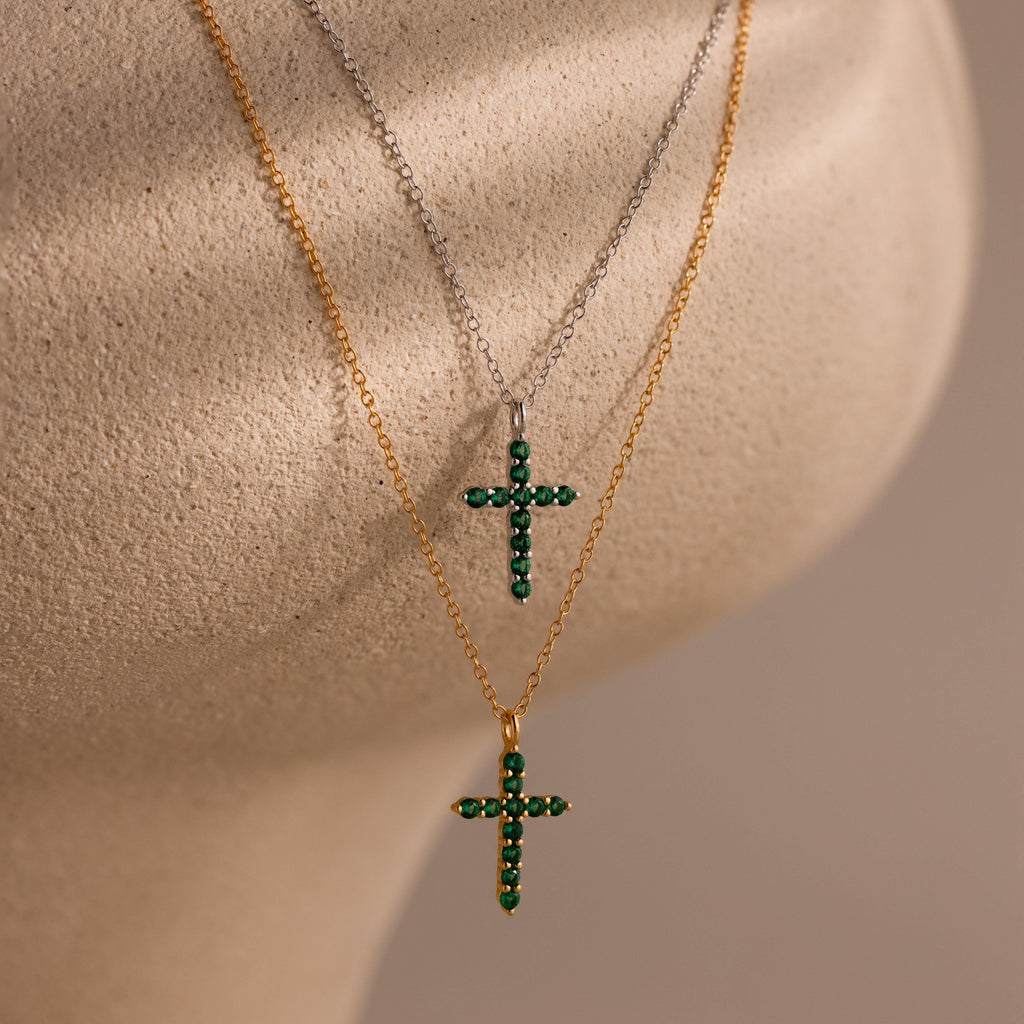 Two gold chain necklaces displayed on a textured beige surface, one with a cross pendant set with round white gemstones, the other with a cross pendant set with round green gemstones.