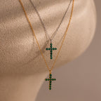 Two gold chain necklaces displayed on a textured beige surface, one with a cross pendant set with round white gemstones, the other with a cross pendant set with round green gemstones.