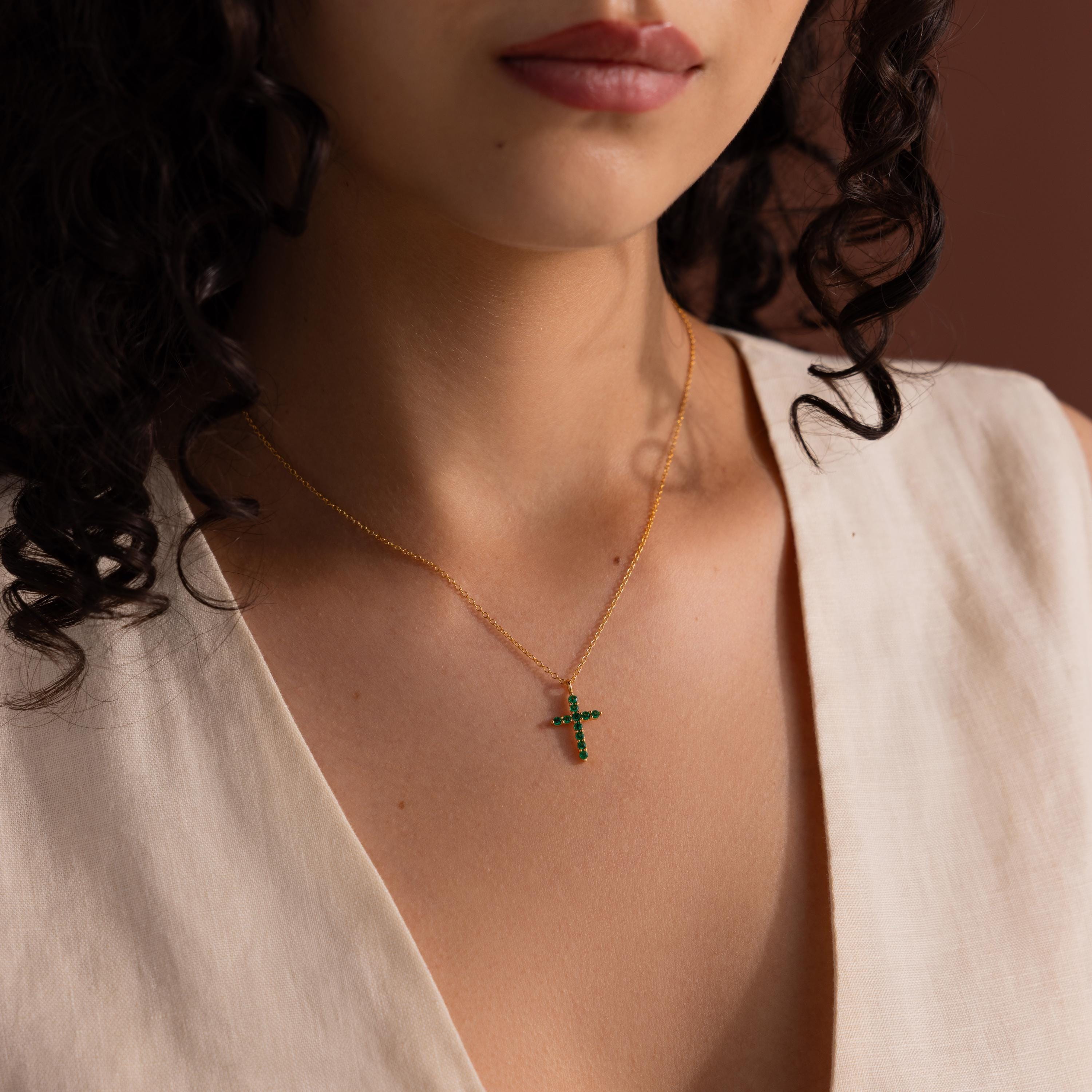 Woman wearing a gold chain necklace with a small cross pendant adorned with green gemstones, paired with a cream sleeveless V-neck top.