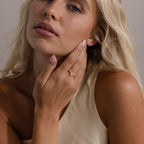 Woman wearing the Radiant Tear Ring in gold with a pear cut diamond center and marquise side stones, styled with glossy lips, nude nails, and a cream one shoulder top.