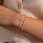 Two gold bracelets worn together, one featuring a delicate cross shaped charm set with small round white gemstones, and the other a tennis bracelet with a row of clear round stones.