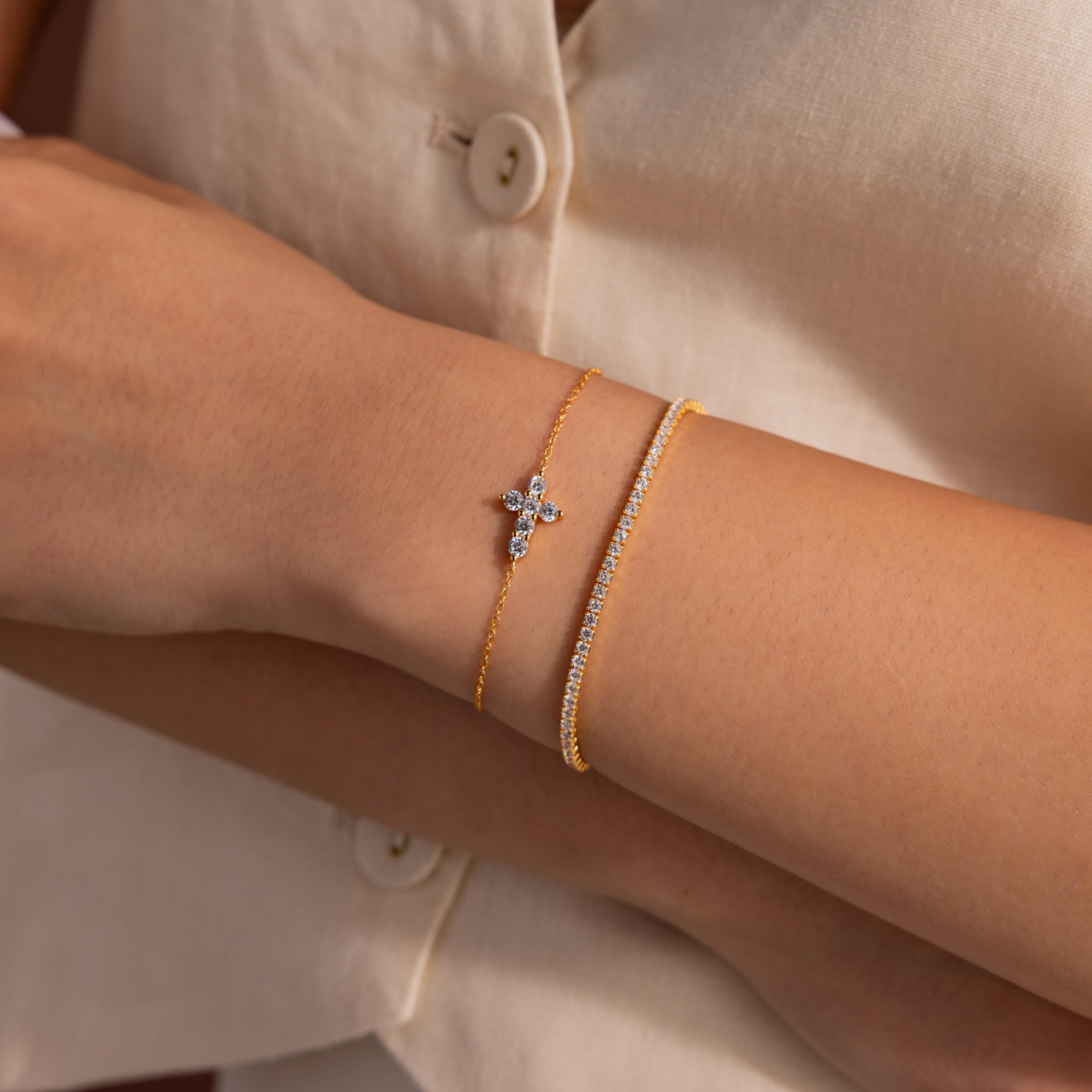 Two gold bracelets worn together, one featuring a delicate cross shaped charm set with small round white gemstones, and the other a tennis bracelet with a row of clear round stones.