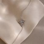 Silver chain bracelet displayed on a smooth, curved ceramic surface, featuring a small cross shaped charm set with round white gemstones.