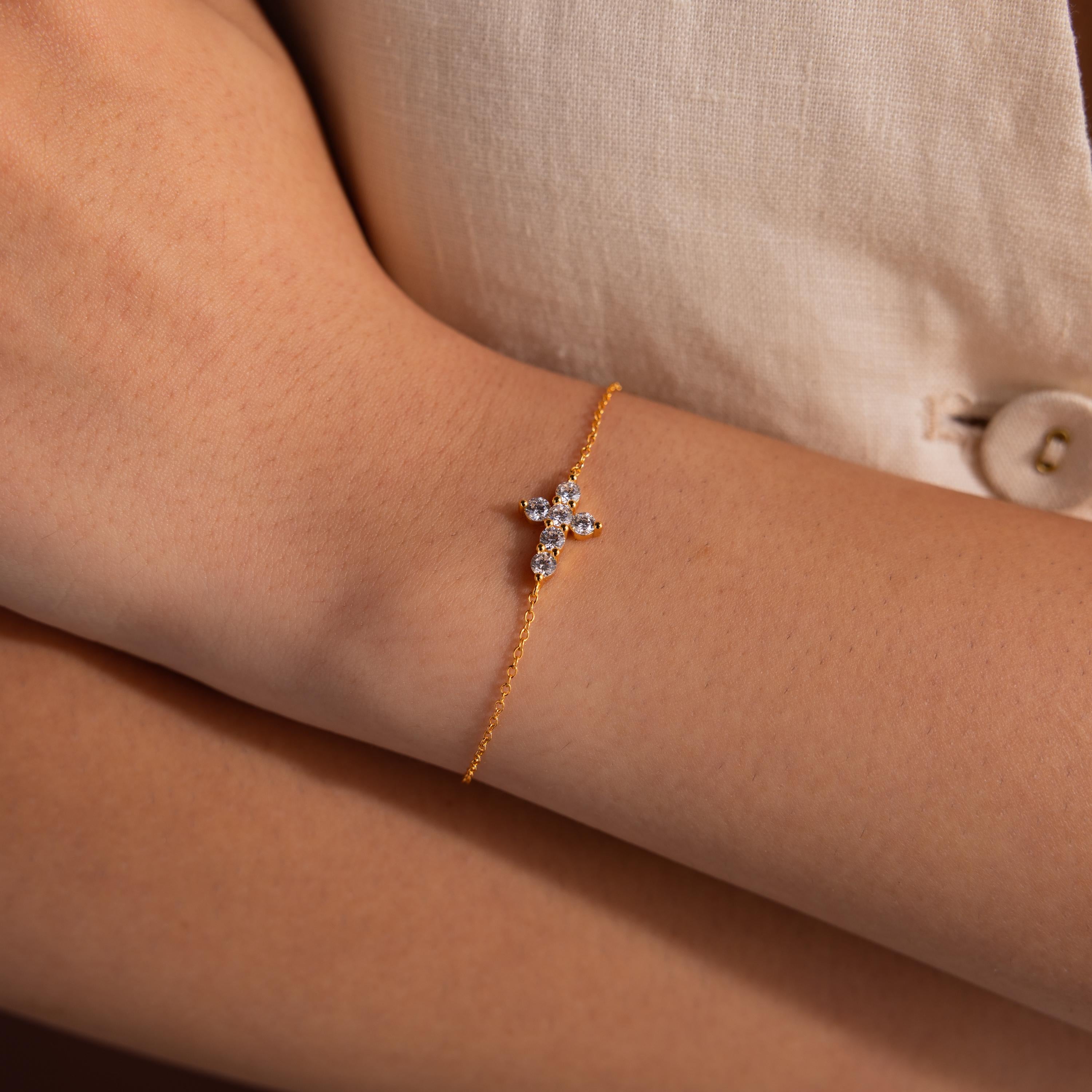 Close-up of a woman's wrist wearing a delicate gold chain bracelet with a small cross shaped charm adorned with round cut white gemstones, paired with a cream fabric background.