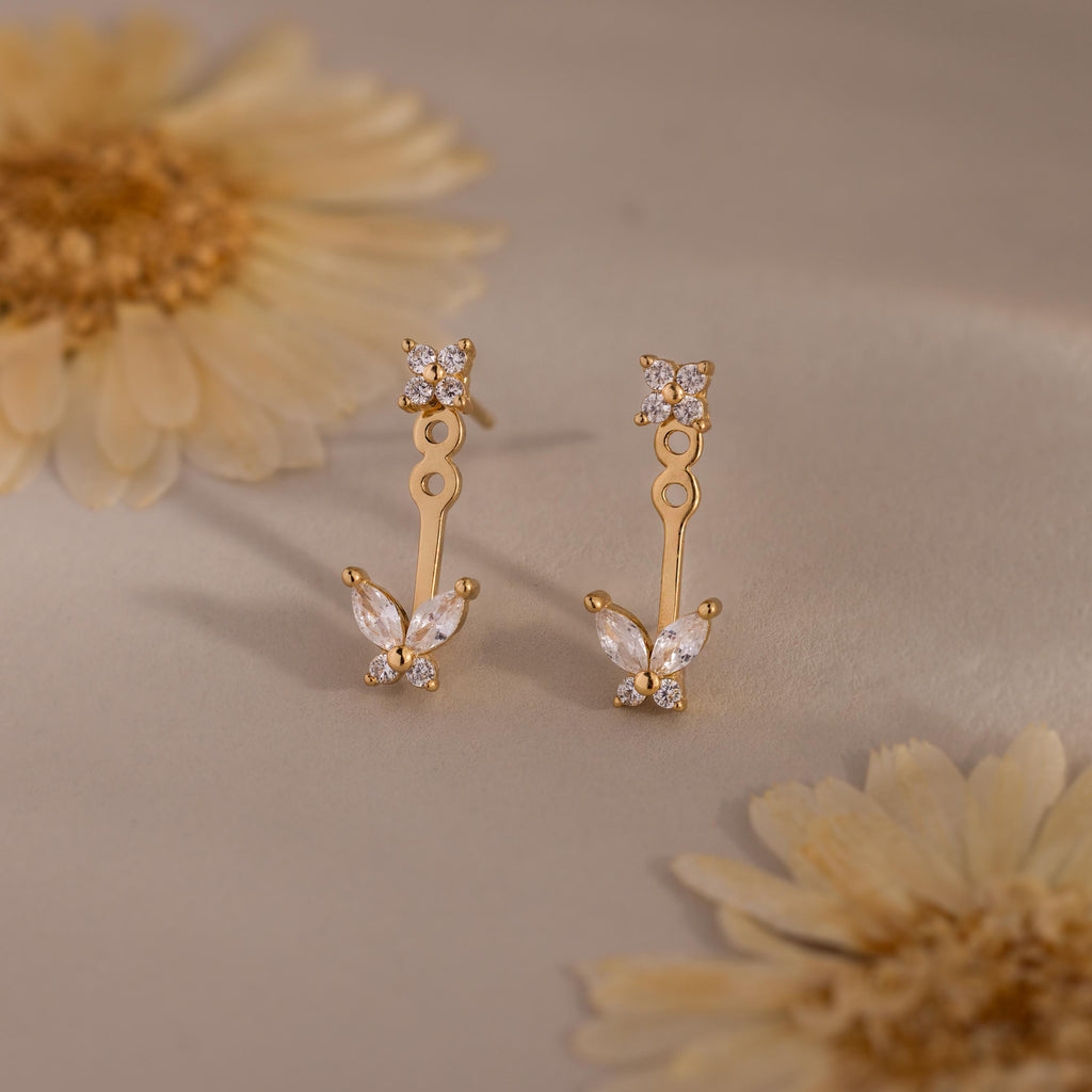Gold stud earrings with diamond clusters and detachable butterfly shaped drops featuring marquise stones, displayed on a soft neutral surface with flowers.