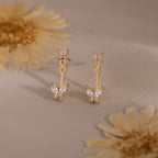 Gold stud earrings with diamond clusters and detachable butterfly shaped drops featuring marquise stones, displayed on a soft neutral surface with flowers.