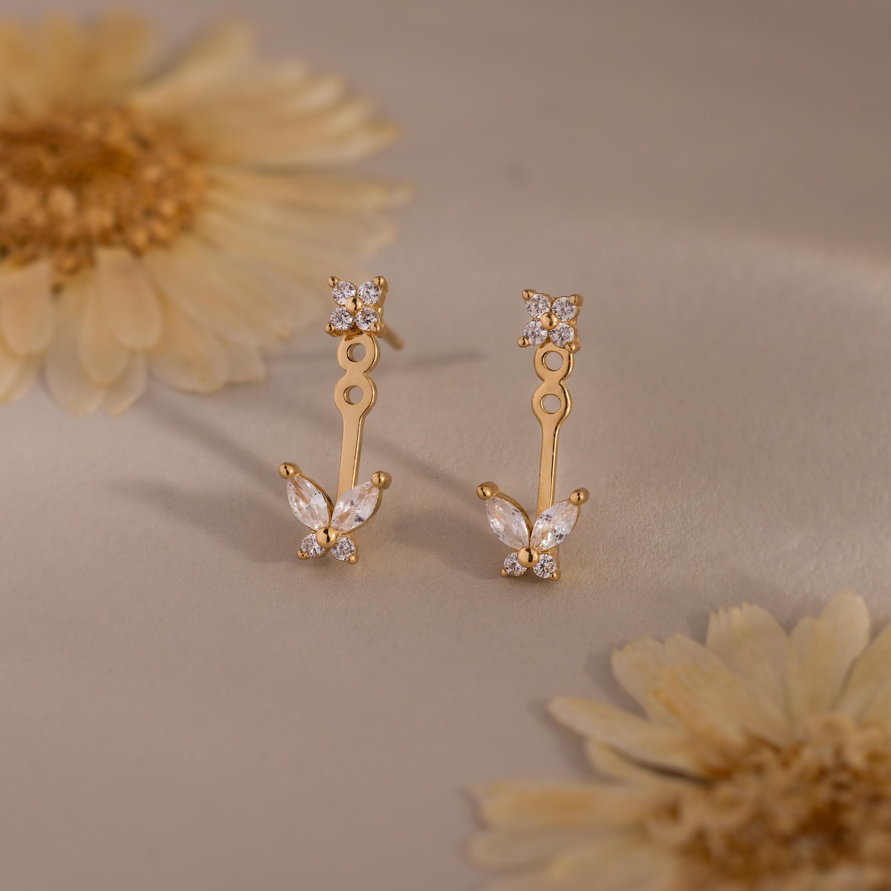 Gold stud earrings with diamond clusters and detachable butterfly shaped drops featuring marquise stones, displayed on a soft neutral surface with flowers.
