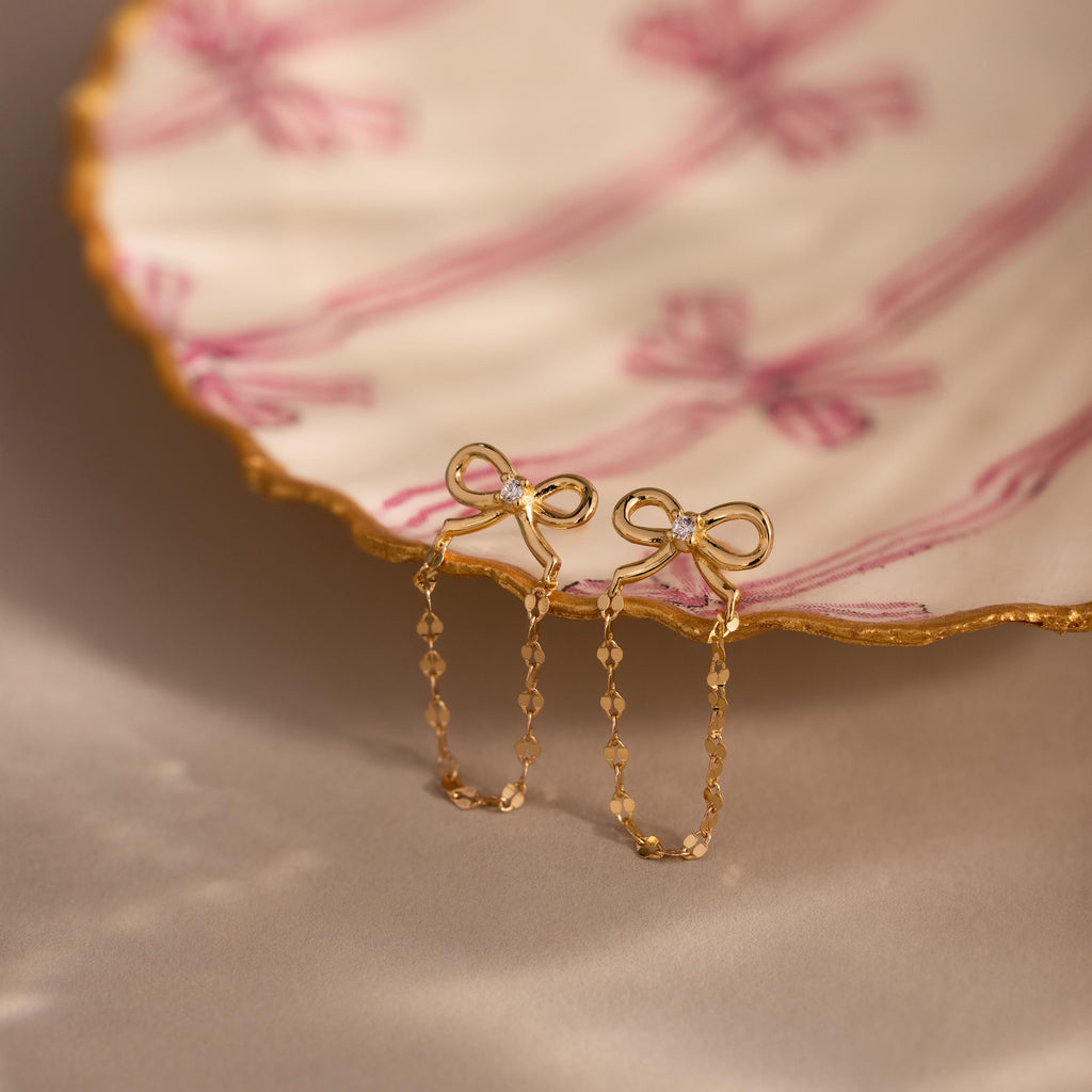 Bow Chain Studs in 18K Gold, featuring delicate chain drops and vintage-inspired design, displayed on a scalloped dish with pink ribbon patterns.