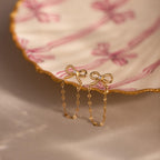 Bow Chain Studs in 18K Gold, featuring delicate chain drops and vintage-inspired design, displayed on a scalloped dish with pink ribbon patterns.
