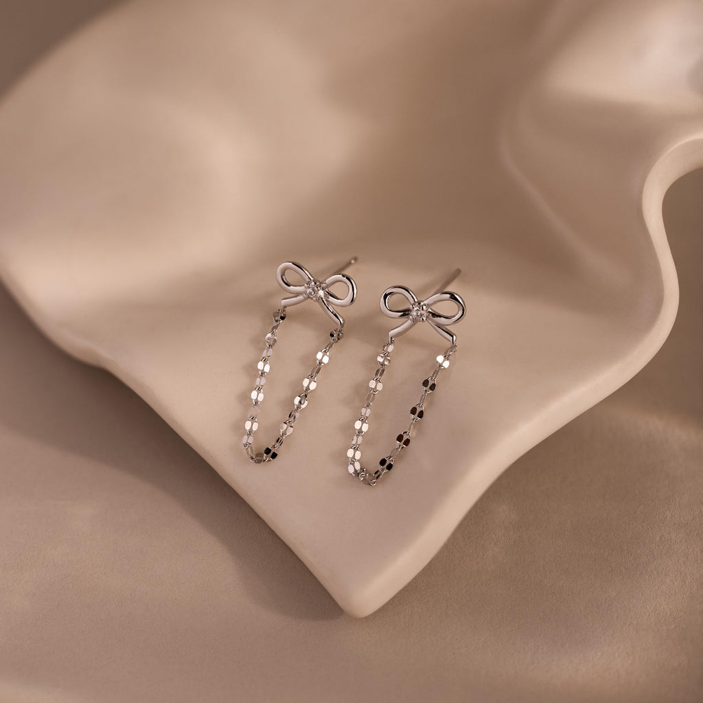 Bow Chain Studs in Sterling Silver are displayed on smooth beige fabric, showcasing their delicate loops and offering a touch of vintage-inspired charm.